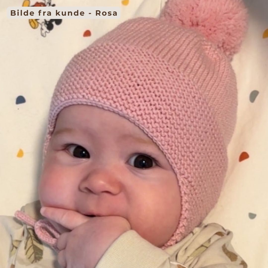 Kundebilde – baby med hjemmestrikket drømmelue i rosa. Strikkepakke drømmelue i 100% kløfri merinoull med dusk, ørelapper og knyttesnor. Den beste lua for små barn – nybegynnervennlig strikkeoppskrift for høst og vinter.