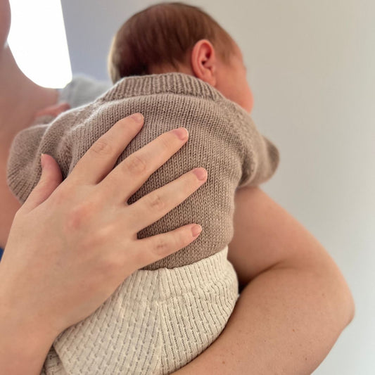 Strikket baby body sett bakfra – ingen knapper i nakke eller hals, kun elastisk hals for lett av- og påkledning. Strikkepakke blomsterbody i 100% merinoull med knapper mellom beina for enkelt bleiebytt. Lettstrikket body – nybegynnervennlig babystrikk.