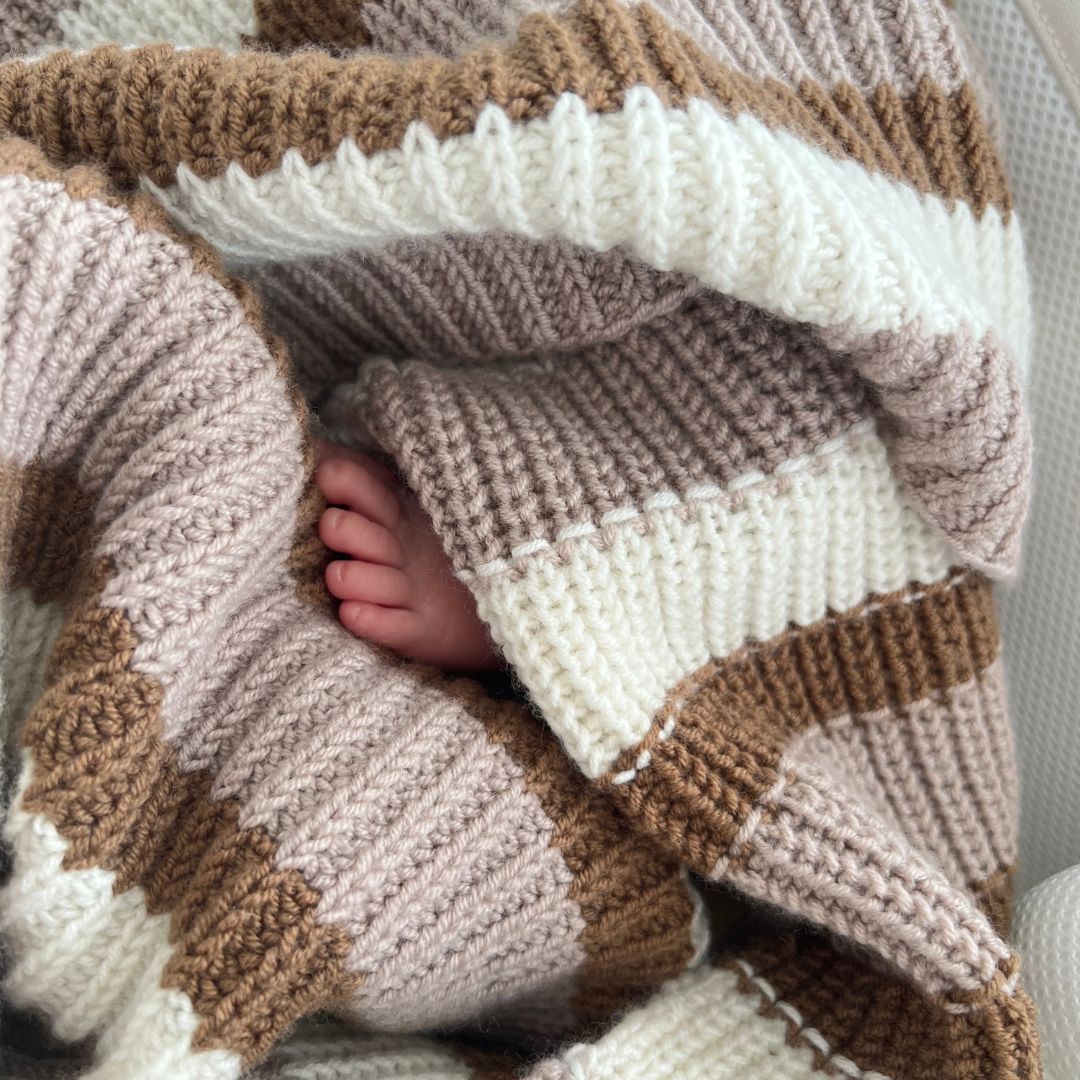 Liten babyfot som gjemmer seg i Vesleskattens yndlingspledd – babyteppe i 100% kløfri merinoull med halvpatent. Nybegynnervennlig strikkepakke babypledd og vognteppe, lett å strikke. Babyens første teppe.