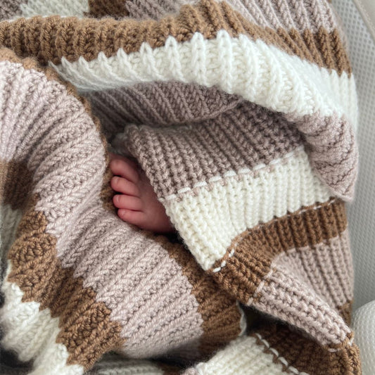 Liten babyfot som gjemmer seg i Vesleskattens yndlingspledd – babyteppe i 100% kløfri merinoull med halvpatent. Nybegynnervennlig strikkepakke babypledd og vognteppe, lett å strikke. Babyens første teppe.