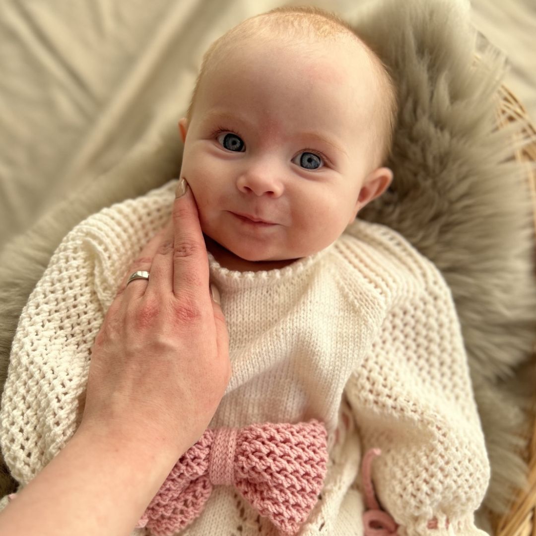 Nærbilde av baby i strikket dåpskjole – strikkepakke dåpskjole med valgfritt silkebånd eller strikket bånd. Strikke dåpskjole til baby uten knapper, med knytting i nakken. Unik og tradisjonell strikkepakke dåpskjole.