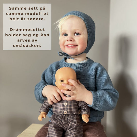 Barn som leker med dukke i Vesleskattens drømmesett – genser og lue i 100% kløfri merinoull med rillemønster. Strikkepakke til barnehagebarn 0–3 år, bra for aktive barn. Godt som eneste ytterplagg på høst og vår, eller under vinterdressen.