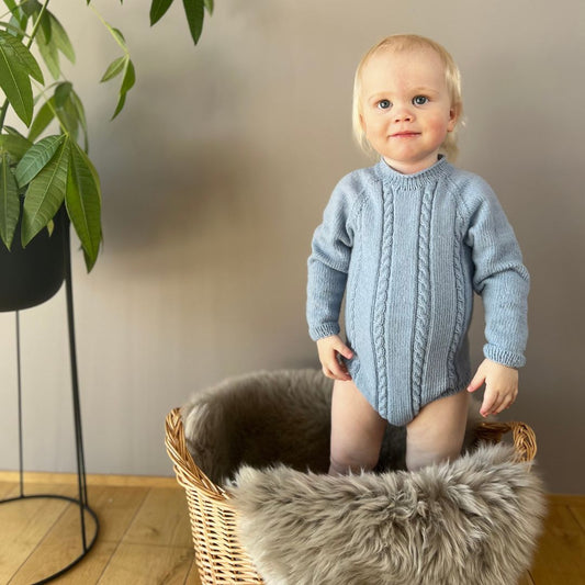 Vesleskattens flettebody forfra – strikkepakke baby i 100% kløfri merinoull med flettemønster. Ingen knapper i halsen, elastisk hals og knapper mellom beina for enkelt bleiebytt. Størrelser fra nyfødt til 2,5 år.