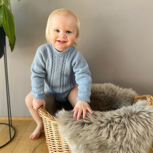 Vesleskattens flettebody på barn – strikkepakke baby i 100% kløfri merinoull med flettemønster. Elastisk hals, ingen knapper i halsen og knapper mellom beina for enkelt bleiebytt. Størrelser fra nyfødt til 2,5 år.
