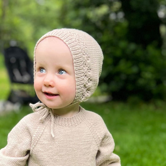 Strikket søt kyse til baby i 100% kløfri merinoull med hullmønster og praktisk knyttesnor – strikkepakke babylue 0–3 år. Fellingen i bakhodet former en dekorativ blomst. Lett å kle av og på, kan brukes året rundt.