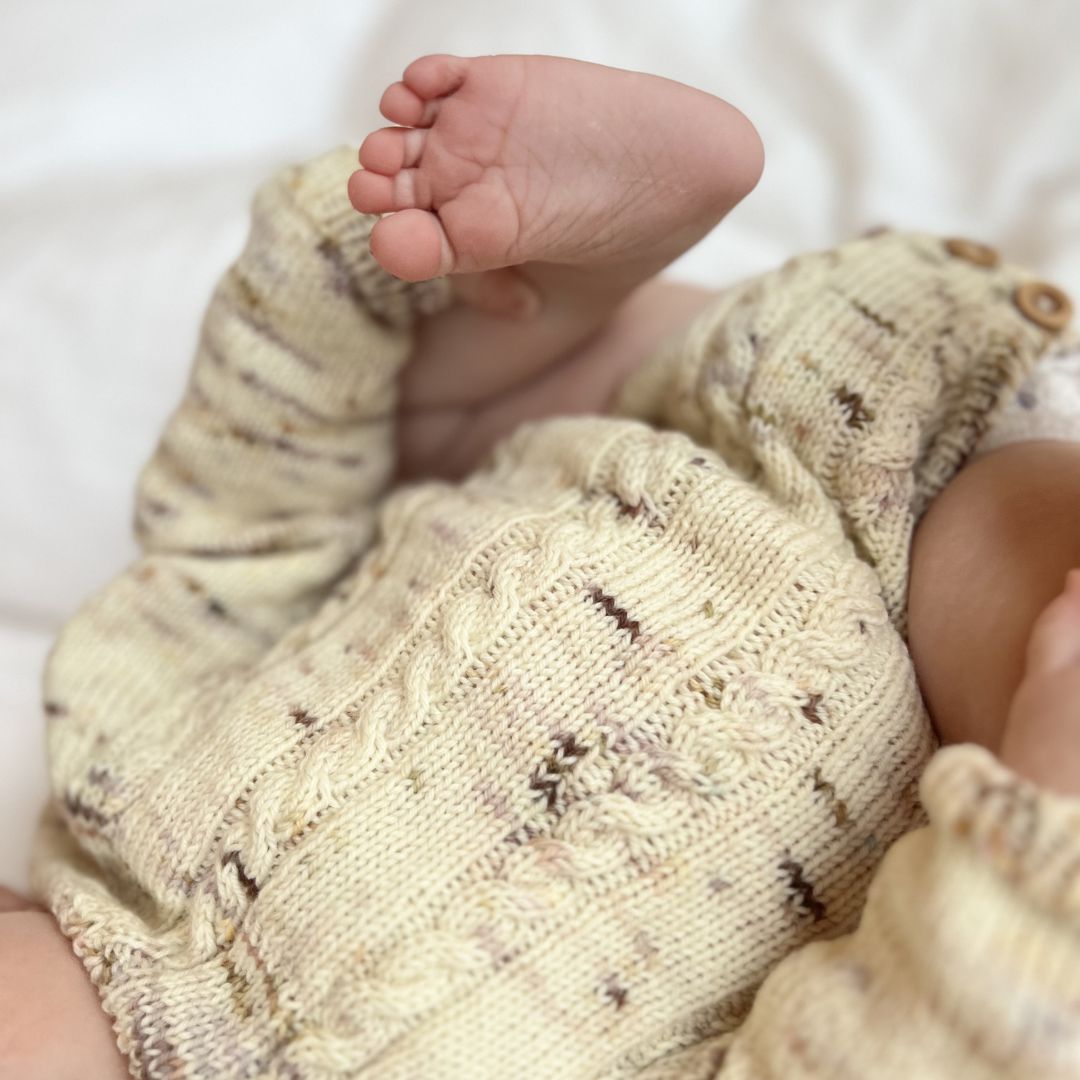 Detaljer forfra på Vesleskattens flettebody – strikkepakke baby i håndfarget eksklusivt Unikt Garn med flettemønster. Størrelser nyfødt til 2,5 år, knapper mellom beina for enkelt bleiebytt.