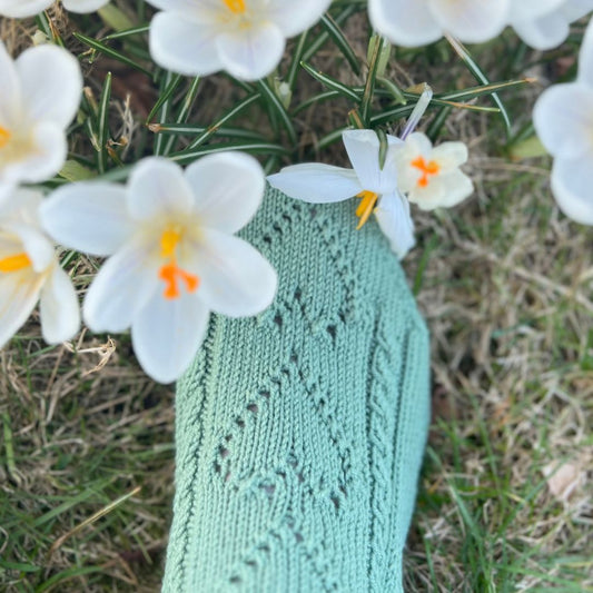 Detaljer av hjertene på Mammas hjertesokker blant blomster ute – nydelig hullmønster som former hjerter i 100% kløfri merinoull. Strikkepakke ankelsokker, perfekt gavetips til å strikke til venninne eller mamma.