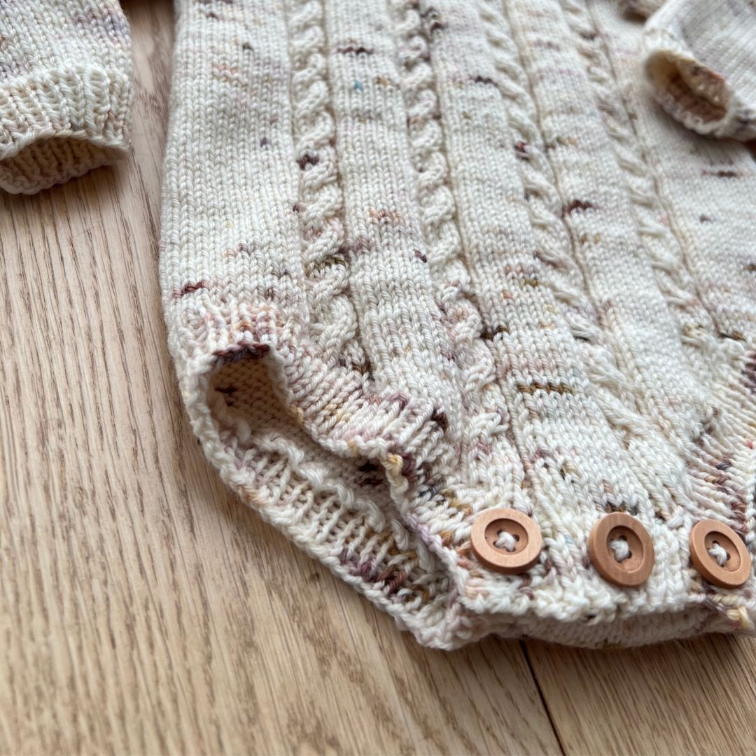 Detaljer på knapper mellom beina på Vesleskattens flettebody – enkelt bleiebytt. Strikkepakke body i håndfarget eksklusivt Unikt Garn med flettemønster, størrelser nyfødt til 2,5 år.