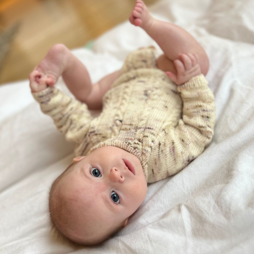 Vesleskattens flettebody ovenfra – baby som leker med føttene. Strikkepakke body i håndfarget eksklusivt Unikt Garn med flettemønster. Størrelser fra nyfødt til 2,5 år, knapper mellom beina for enkelt bleiebytt.
