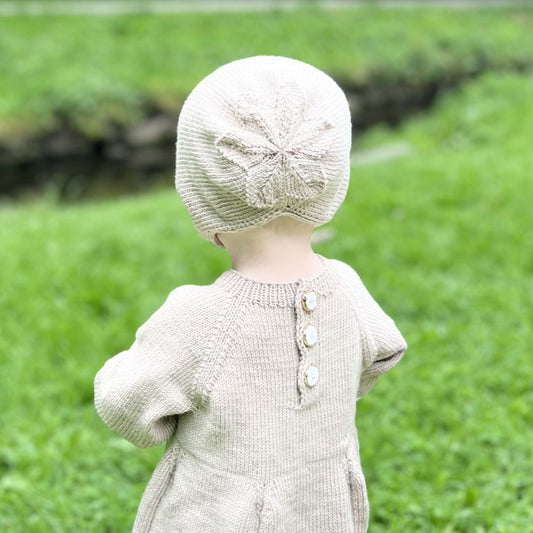 Søt kyse sett bakfra – fellingen i bakhodet former en dekorativ blomst. Strikket babylue i 100% kløfri merinoull med hullmønster og knyttesnor – strikkepakke 0–3 år. Kan brukes året rundt.