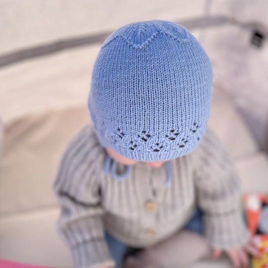 Søt kyse sett ovenfra – blomsterfelling i bakhodet som former en dekorativ blomst. Strikket babylue i 100% kløfri merinoull med hullmønster – strikkepakke kyse 0–3 år. Kan brukes året rundt.